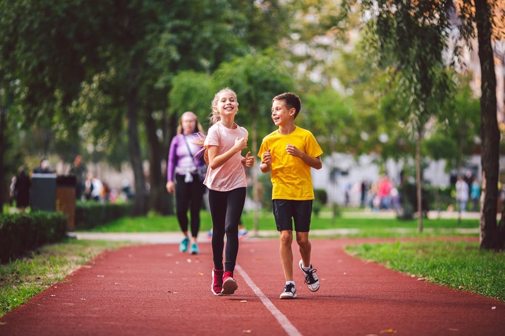 Two children jog together on a red track in a park, smiling and chatting, with other people walking in the background - Tobin C Gallawa DPM FACFAS Two children jog together on a red track in a park, smiling and chatting, with other people walking in the background - Tobin C Gallawa DPM FACFAS